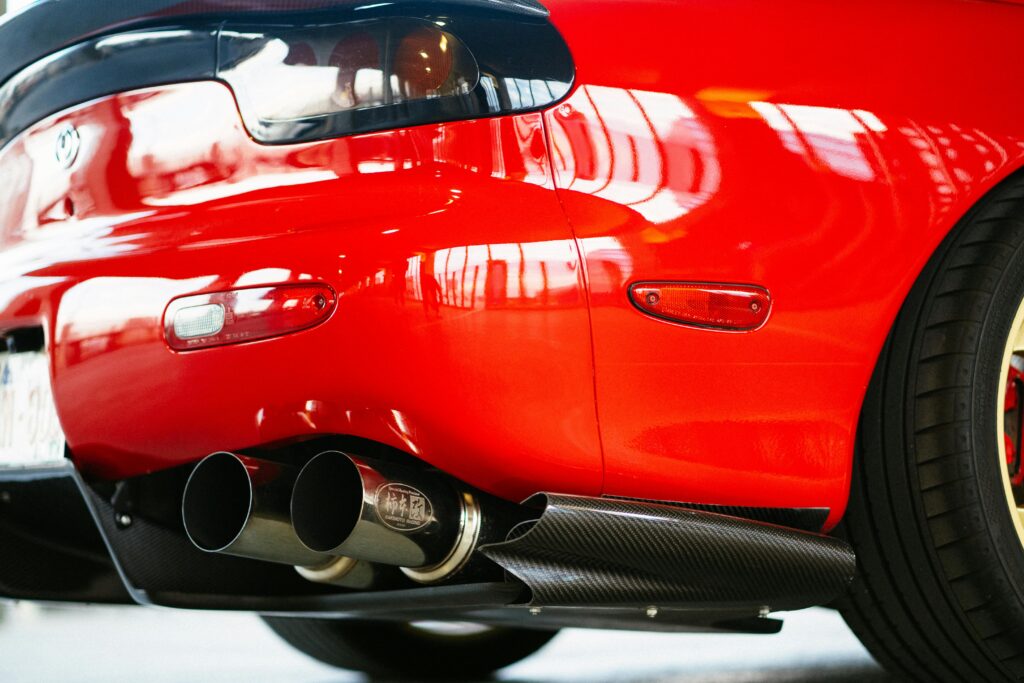 Rear close-up of a modified red Mazda RX-7 (FD) featuring a carbon fiber rear diffuser, a dual-tip Kakimoto Racing exhaust system, and signature sleek curved bodywork