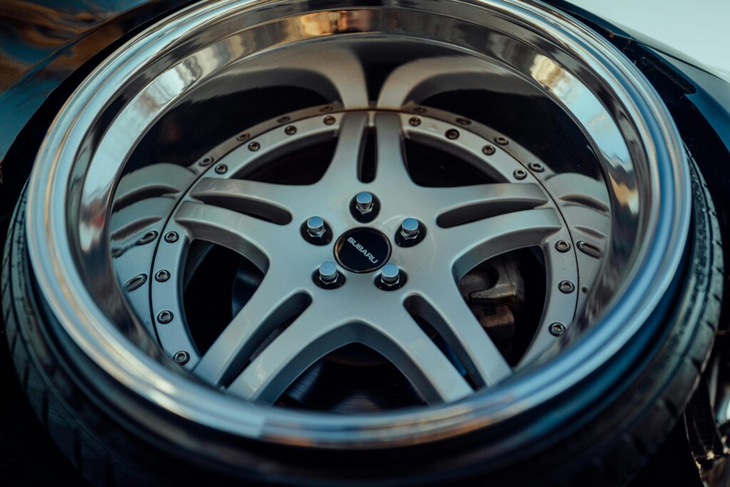 A close-up, low-angle shot of a custom deep-dish multi-piece alloy wheel on a Subaru. The wheel features a classic five-spoke star design in a matte silver finish, secured with visible perimeter hardware bolts and a black Subaru center cap