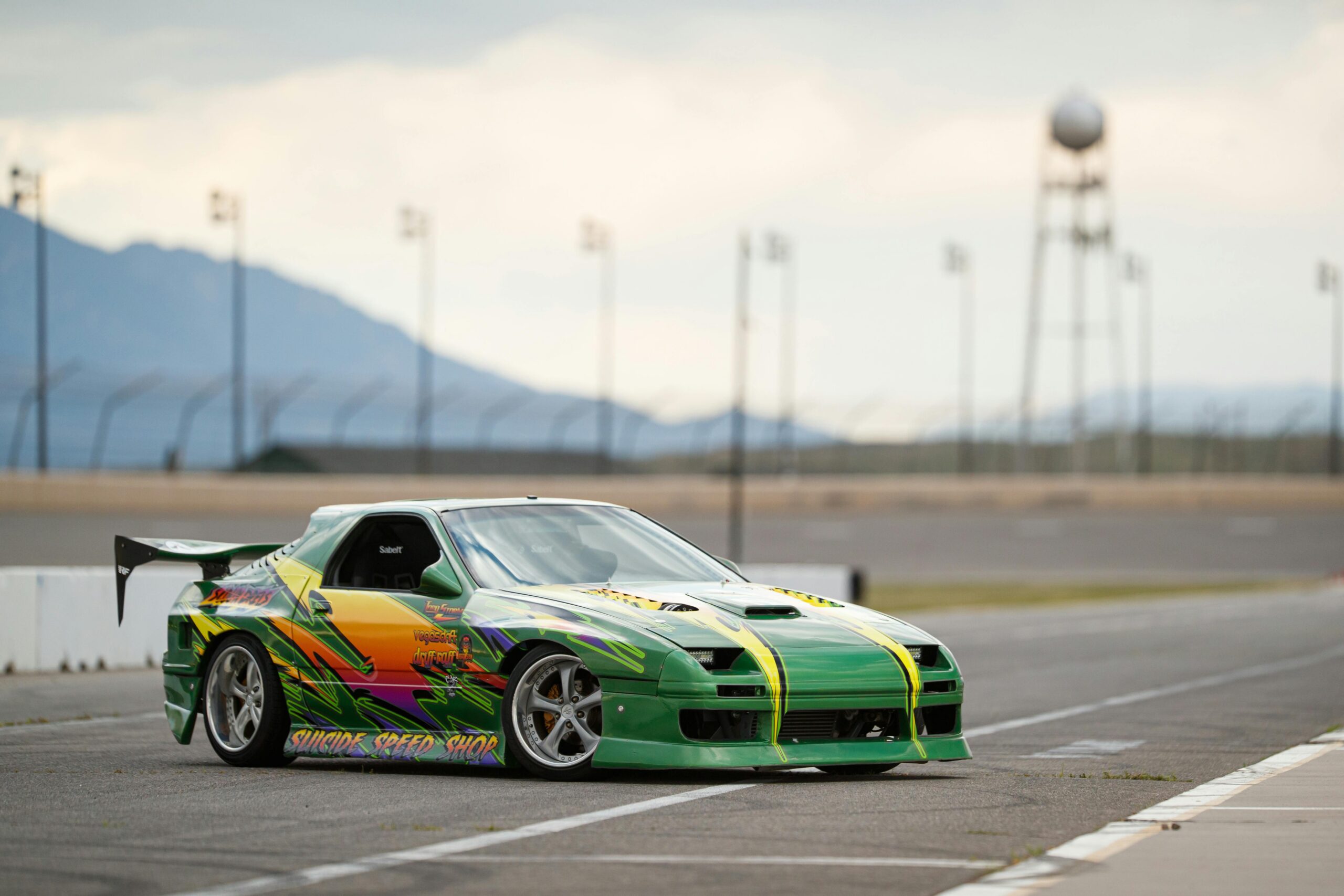 A modified green Mazda RX-7 (FC) drift car on a race track, featuring a widebody kit, large rear wing, and vibrant multi-coloured livery decals